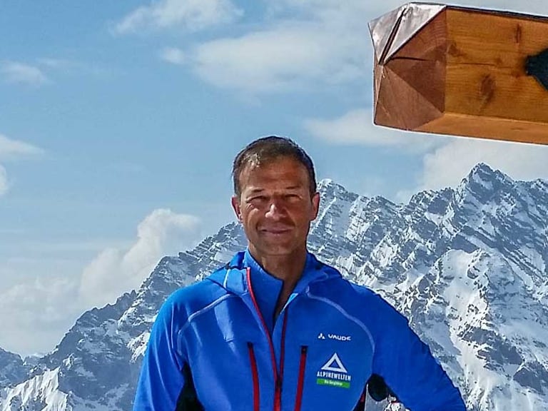 Am hölzernen Gipfelkreuz steht Bergführer Michael Höglauer in blauer Softshell-Jacke vor Panorama schneebedeckter Berggipfel.