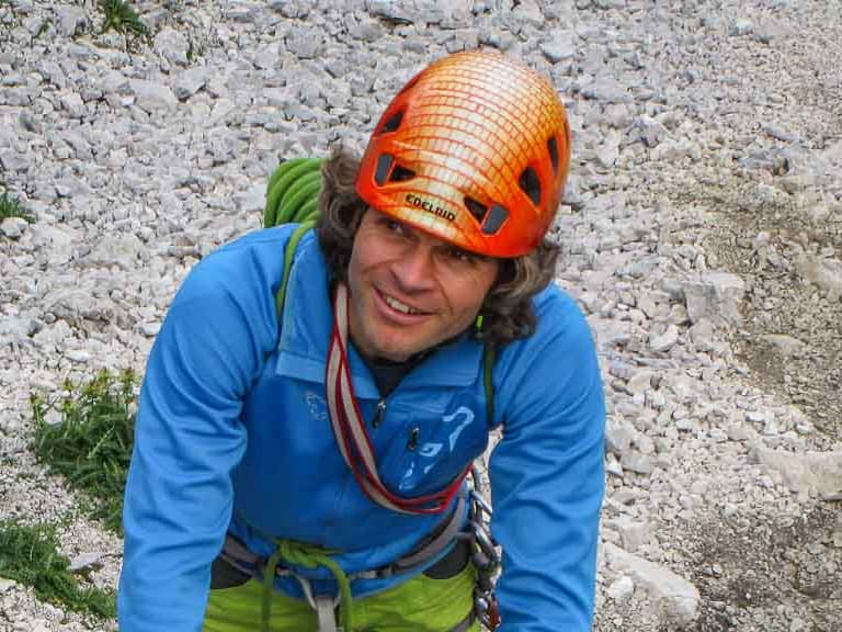 Bergführer Michael Hornsteiner in orangem Helm klettert an Stahlleiter eines Klettersteigs, gesichert am Drahtseil über Geröllfeld.
