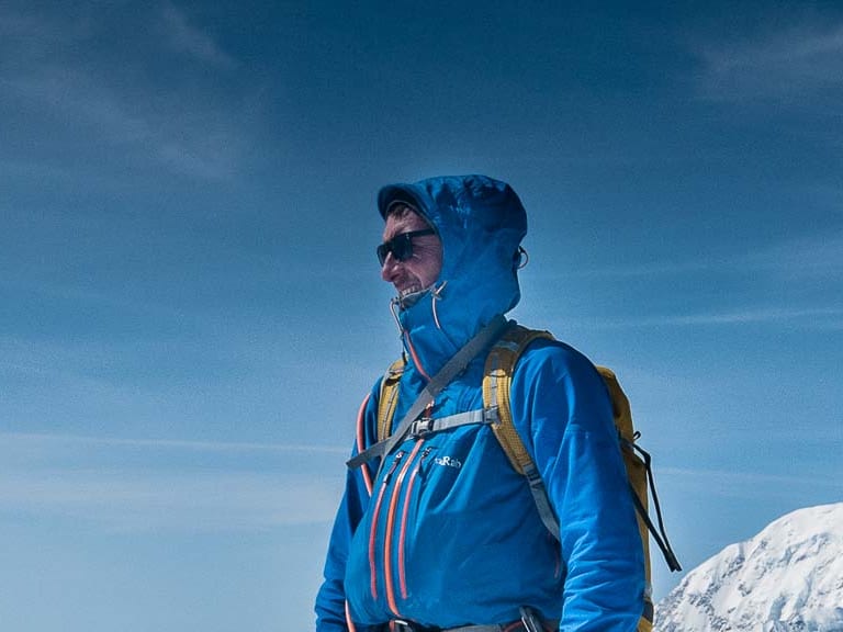 Bergführer Christoph Hummel steht in blauer Bergsteigerausrüstung auf verschneitem Gipfel vor schneebedeckter Hochgebirgslandschaft.
