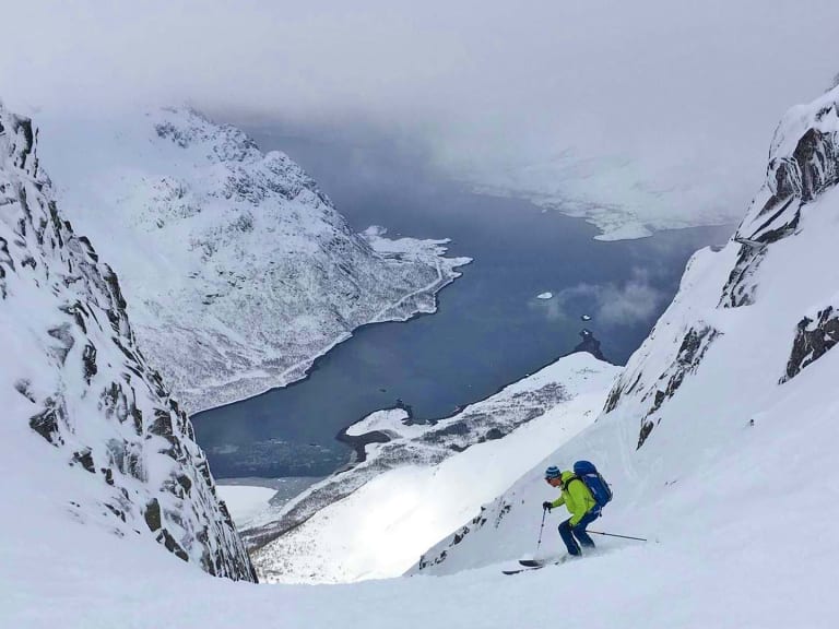 Bergführer Simon Kraus fährt auf Skiern einen steilen Hang hinab mit tiefblauem Fjord und Bergen im Hintergrund.