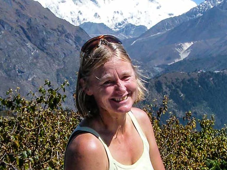 Portrait von Bergwanderführerin Isolde Fink im weißen Top vor schneebedeckten Berggipfeln und grüner Vegetation.