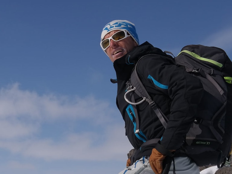 Auf verschneitem Felsvorsprung steht Bergführer Jörg Rauschenberger mit blauer Mütze und schwarzer Softshell-Jacke.