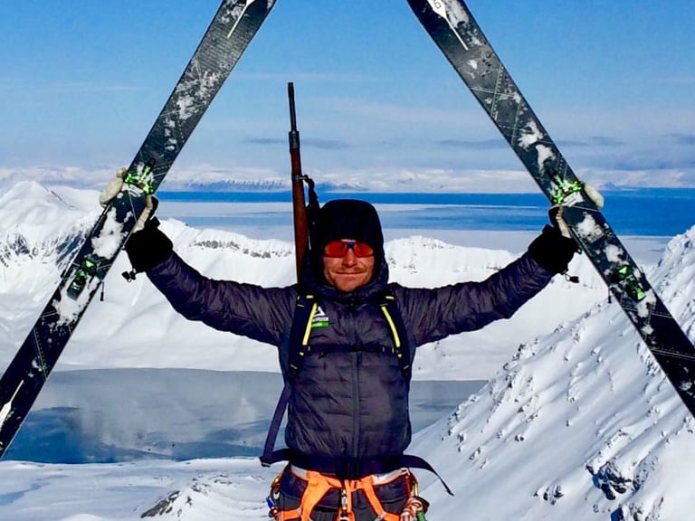 Bergführer Marcel Roßbach hält seine Skier triumphierend über den Kopf auf einem verschneiten Gipfel.