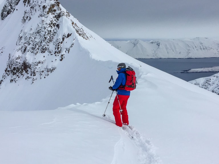 Bergführer Florian Schmidt in roter Hose und blauer Jacke steigt einen verschneiten Hang in isländischer Fjordlandschaft auf.