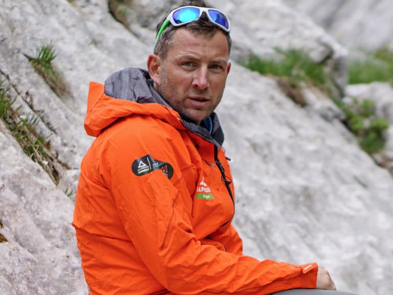 Guide Markus Schubert sitzt in orangefarbener Outdoorjacke auf hellem Kalksteinfels mit Klettergurt und Bergschuhen.