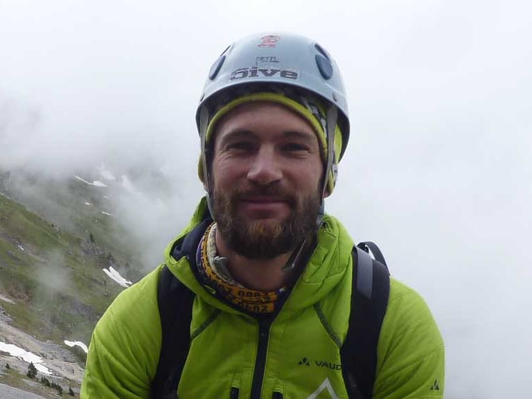 Bergführer Philipp Berg trägt weißen Kletterhelm und schwarzen Klettergurt vor alpiner Berglandschaft mit Kalksteinfelsen.