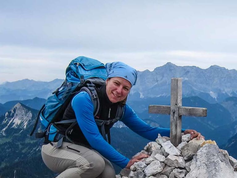 Bergwanderführerin Sandra Wolf kniet neben einem Steinmännchen mit Holzkreuz auf einem Berggipfel und lächelt in die Kamera.