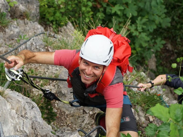 Bergführer Christian Knabe klettert am Drahtseil einer Felswand in roter Jacke und weißem Helm mit Klettersteig-Set.