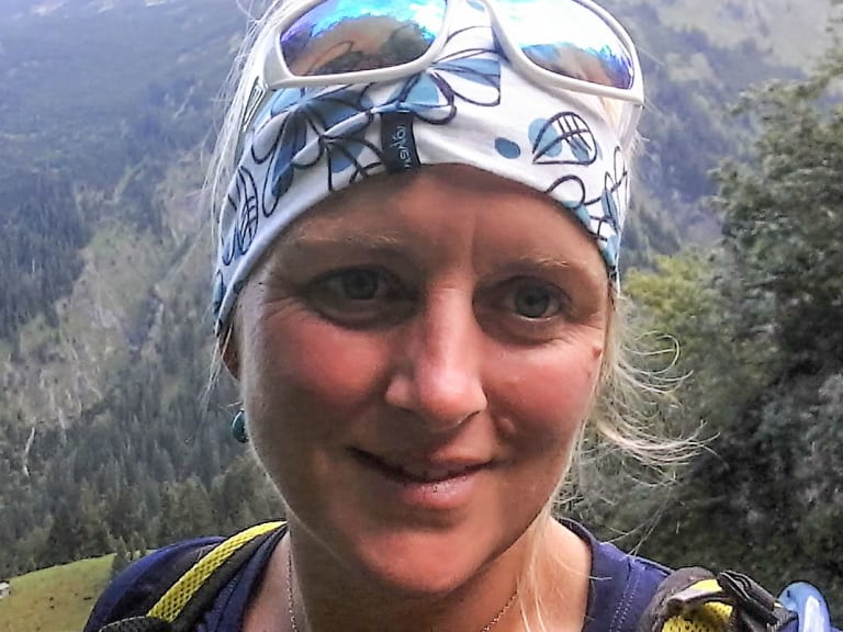 Bergwanderführerin Maria Anna Huber lächelt mit weißer Gletscherbrille und blauem Multifunktionstuch vor bewaldeter Berglandschaft.