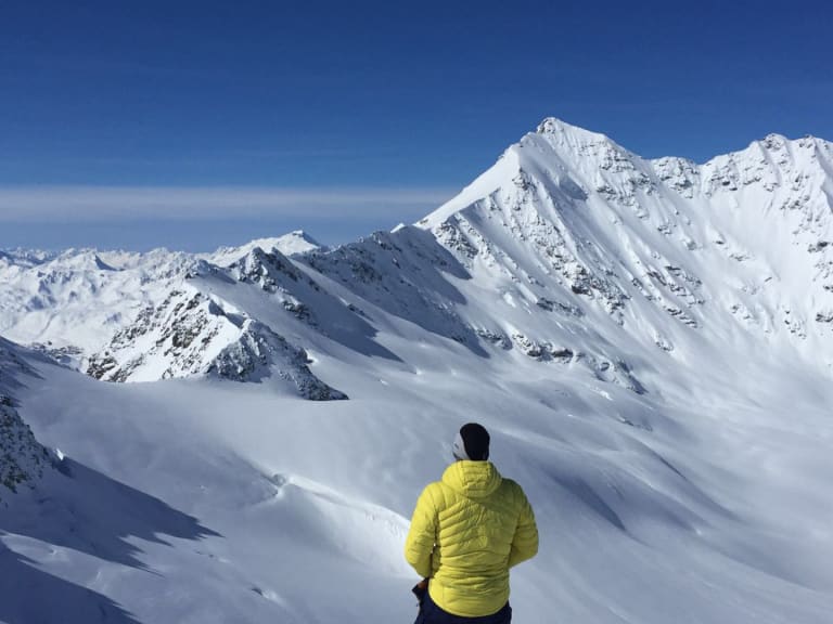 Bergführer Rene Hellmann in gelber Daunenjacke betrachtet schneebedeckte Gipfel am Elbrus bei strahlendem Himmel.
