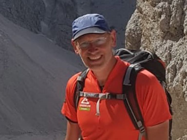 Bergwanderführer Manfred Beier lächelt in blauer Kappe und rotem T-Shirt mit Klettergurt vor hellen Kalksteinformationen.