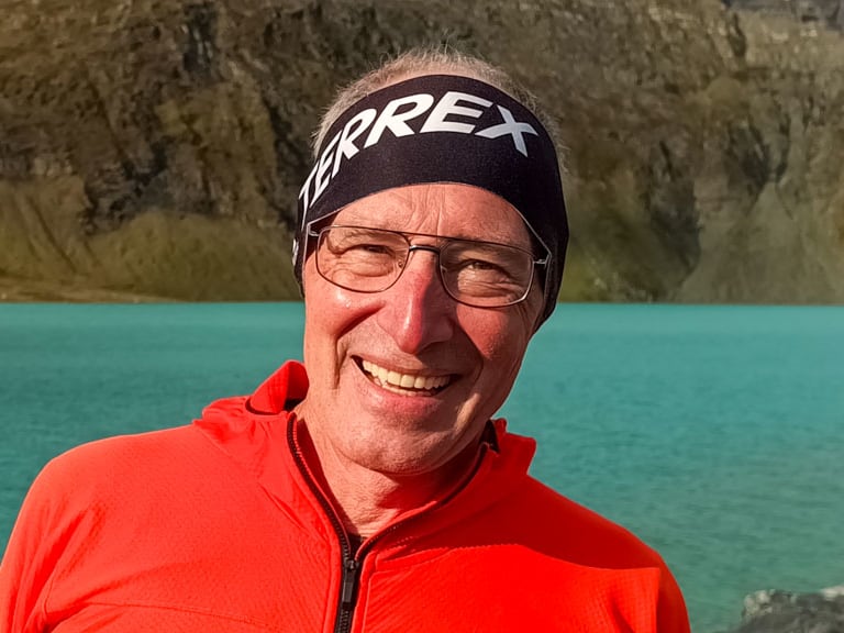 Bergwanderführer Manfred Beier in roter Softshelljacke mit Alpine Welten Logo steht am steinigen Ufer eines türkisfarbenen Bergsees in Norwegen.