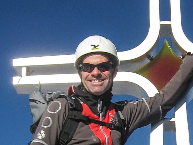 Portrait von Bergwanderführer Andreas Schneider mit weißem Helm und Sonnenbrille vor einem weißen Gipfelkreuz unter blauem Himmel.