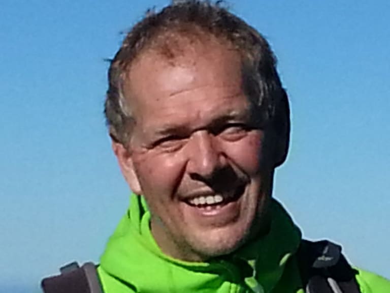 Bergwanderführer Martin Werner lächelt in grüner Bergsportjacke mit Zugspitze-Logo vor blauem Himmel und warmen Berghängen.