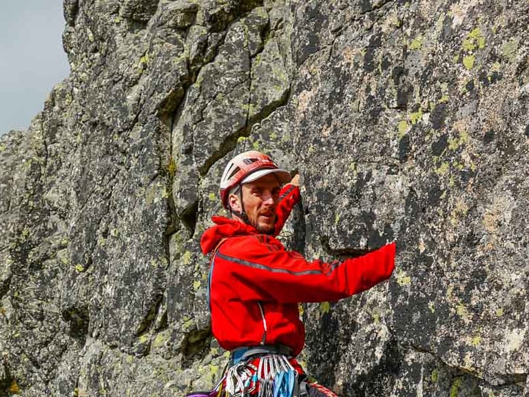 An einer rissigen Felswand mit gelblichen Flechten klettert Bergführer Peter Klug in roter Softshelljacke und rotem Helm.