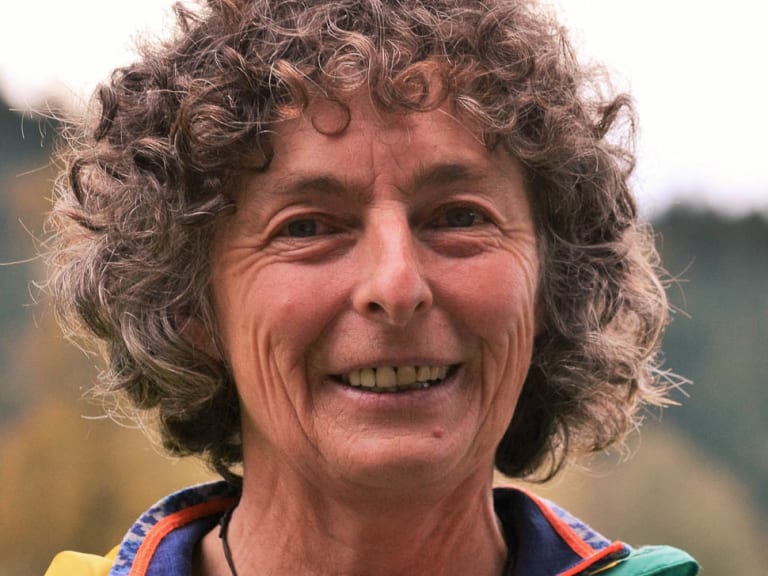 Freundliches Portrait von Bergwanderführerin Irene Aschauer mit lockigem Haar und türkiser Outdoorjacke vor Berglandschaft.