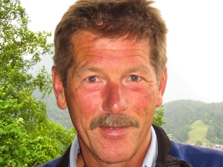 Bergführer Georg Gruber blickt in die Kamera und trägt eine dunkelblaue Jacke, im Hintergrund zeigt sich grüne bewaldete Hügellandschaft.