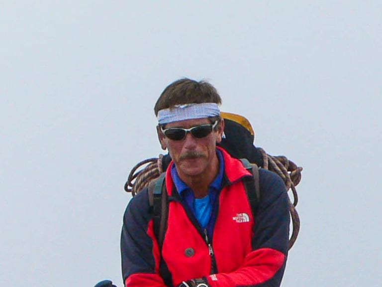 Bergführer Georg Gruber steht mit Wanderstöcken und Rucksack neben einem Metallkreuz auf einem felsigen Gipfel.