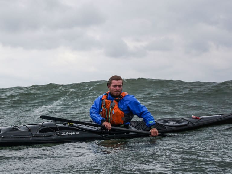 Bergwanderführer Josef Aschauer paddelt in schwarzem Seekajak auf bewegtem Wasser mit Doppelpaddel, trägt blaue Jacke und orange Schwimmweste.
