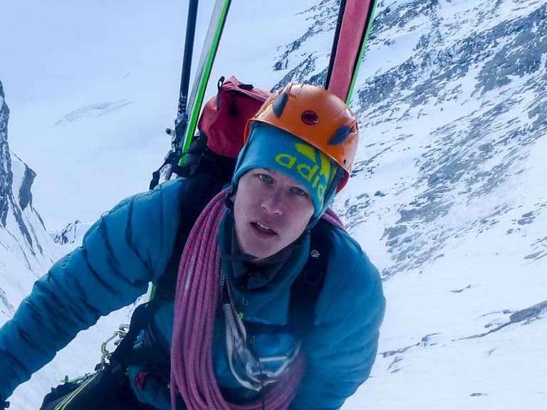 Bergführer Martin Schidlowski kniet mit Eispickel in blauer Daunenjacke im Neuschnee, zwei Skier stehen neben ihm im Schnee.