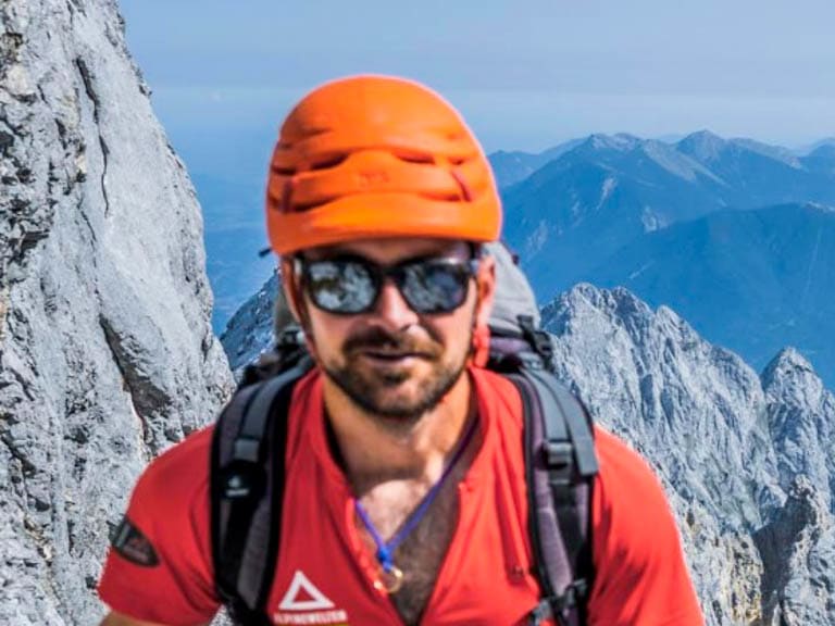 In orangem Helm und rotem Shirt mit schwarzem Klettergurt steht Bergführer Franz Wagner auf hellem Kalkstein vor Felswand.