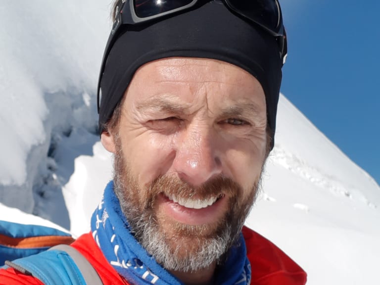 Portrait von Bergführer Michael Aigner mit Gletscherbrille und grauem Bart am Berner Dreigestirn bei strahlendem Sonnenschein.