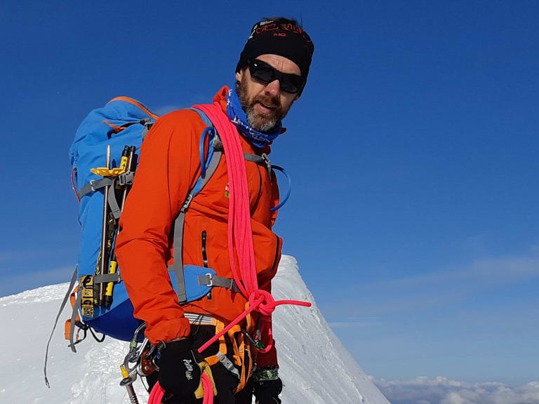 Bergführer Michael Aigner steht mit Steigeisen und orangener Jacke auf schmalem, schneebedecktem Gipfelgrat beim Aufstieg zum Piz Bernina.