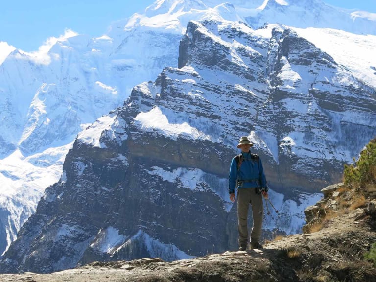 Bergwanderführer Werner Neubauer steht in blauer Jacke und braunem Hut auf steinigem Felsplateau vor schneebedeckten Gipfeln.