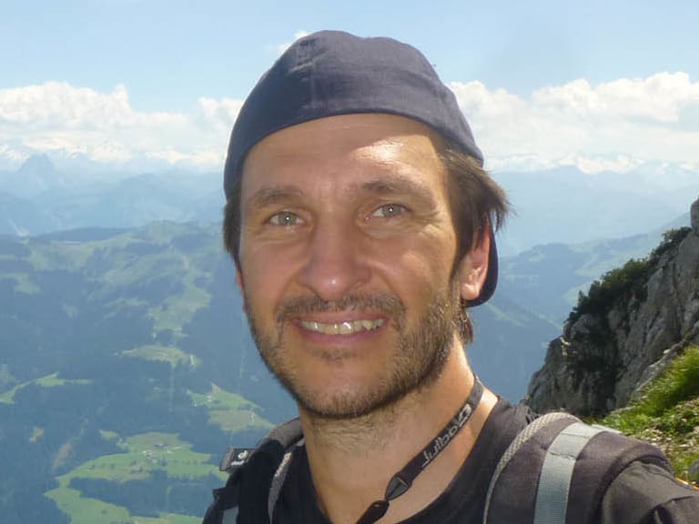 Bergwanderführer Andreas Maier trägt orangefarbene Kletterausrüstung und grauen Rucksack vor Alpenpanorama.