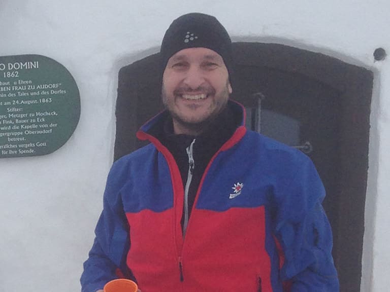 Bergwanderführer Andreas Maier macht Pause vor einer Berghütte mit orangefarbener Tasse in winterlicher Landschaft.