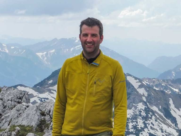 Auf einem Berggipfel im Nationalpark Berchtesgaden lächelt Bergwanderführer Michael Lichtenstern in gelber Jacke.