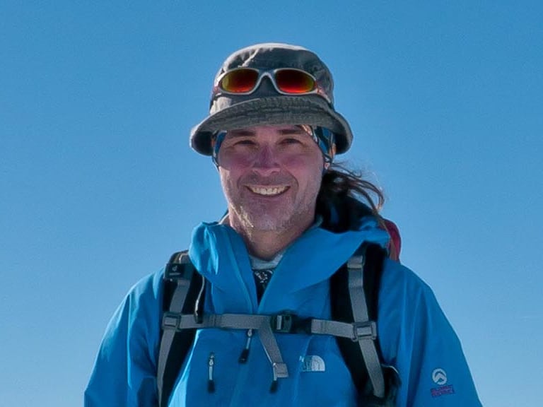 Bergwanderführer Markus Arndt in blauer Softshell-Jacke lächelt auf einem schneebedeckten Gipfel bei Sonnenschein.