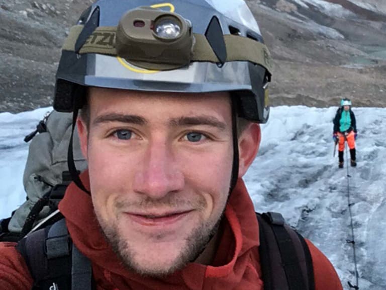 Auf dem Gletscher macht Bergführer Julian Conrad ein Selfie mit Helm und Stirnlampe, eine zweite Person steht mit Skiern dahinter.