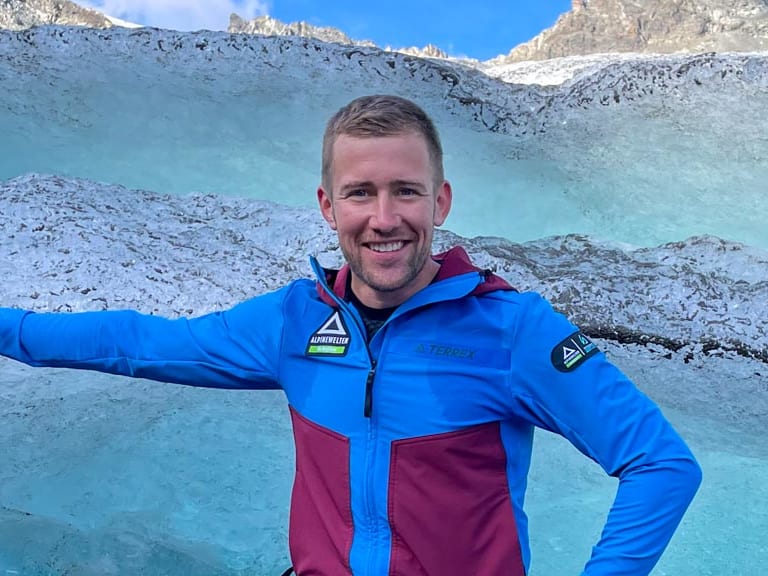 Bergführer Julian Conrad steht in blau-roter Outdoor-Bekleidung präsentierend vor einer Gletscherspalte mit türkisblauen Eiswänden.