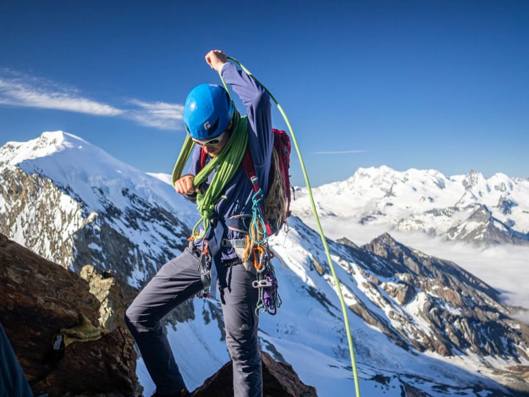 Bergführer Arne Aerts steht auf dunklem Fels und hantiert mit grünem Seil, schneebedeckte Gipfel im Hintergrund.