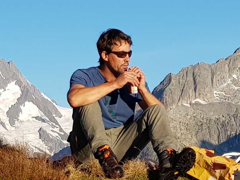 Bergführer Georg Schwarzenberger macht Pause auf grasigem Berghang mit gelbem Rucksack und trinkt aus einer Flasche.