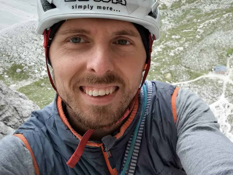 Am hellen Kalkstein lächelt Bergführer Paul Lohse mit weißem Helm und bunten Kletterseilen um den Hals in die Kamera.