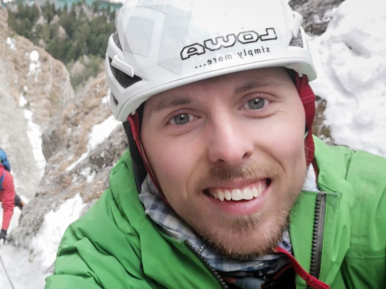 Im Vordergrund lächelt Bergführer Paul Lohse in die Kamera, während eine Person an der schneebedeckten Felswand klettert.