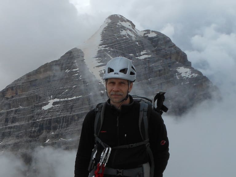Bergwanderführer Thomas Schill in schwarzer Kleidung und weißem Kletterhelm steht auf felsigem Grat vor verschneiter Berglandschaft.