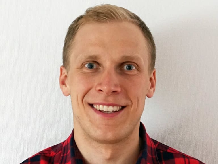 Bergführer Jan Schneider lächelt vor weißer Wand und trägt ein rot-schwarz kariertes Hemd mit Kragen.