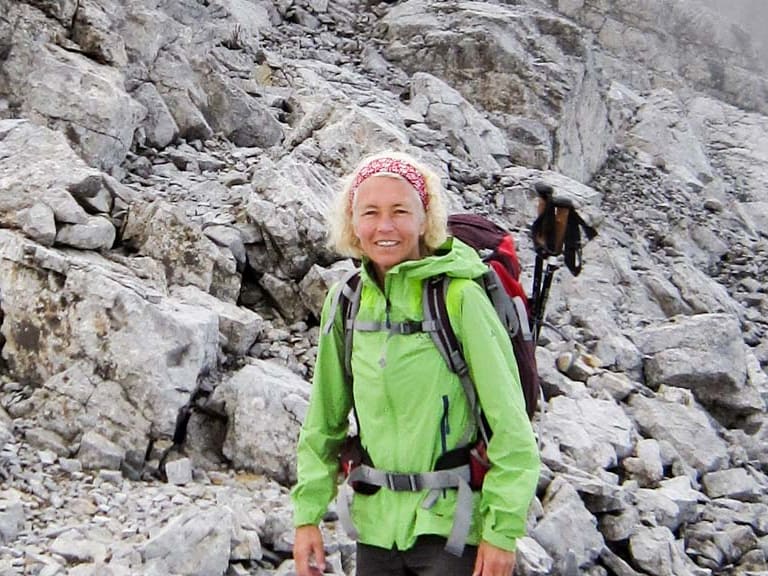 Bergwanderführerin Friedlinde Hillmann steht lächelnd zwischen Kalksteinblöcken in grüner Softshelljacke und rotem Rucksack.
