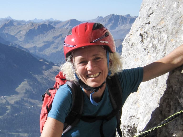 Bergwanderführerin Friedlinde Hillmann klettert mit Klettergurt und rotem Helm an steiler Kalksteinwand über dem Tal.