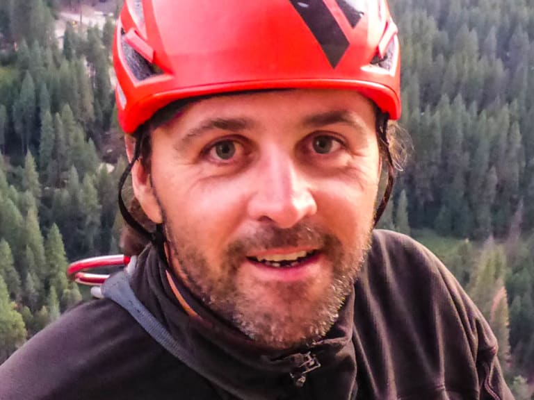 Bergführer Markus Hofmann macht ein Selfie mit rotem Helm am Gruenstein Klettersteig mit Waldblick im Hintergrund.