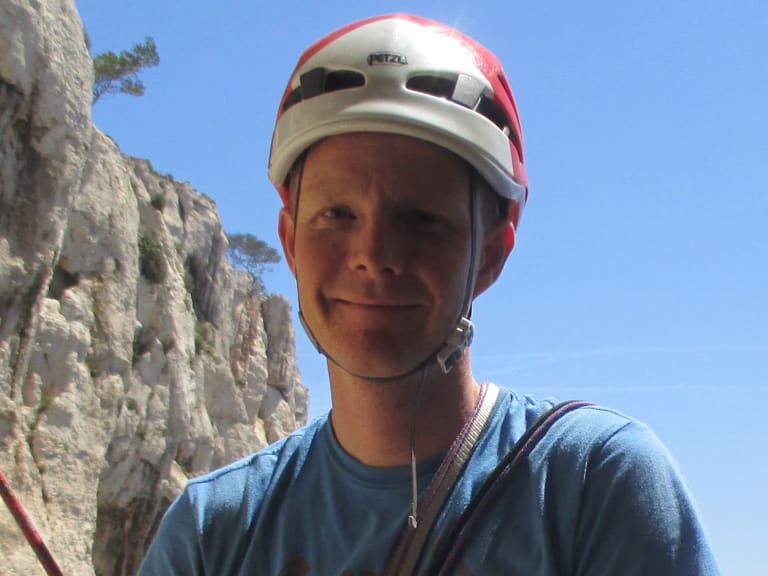 Bergführer Thomas Hochrein trägt rot-weißen Helm und blaues T-Shirt vor Kalksteinwand mit blauem Meer im Hintergrund.