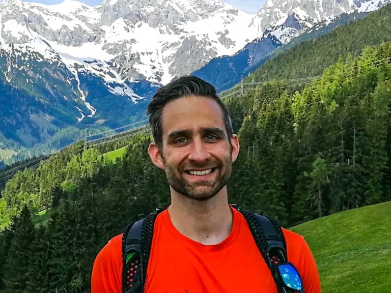 Bergwanderführer Alexander Plotzki lächelt vor schneebedeckten Felsgipfeln und trägt rotes T-Shirt mit schwarzem Rucksack.