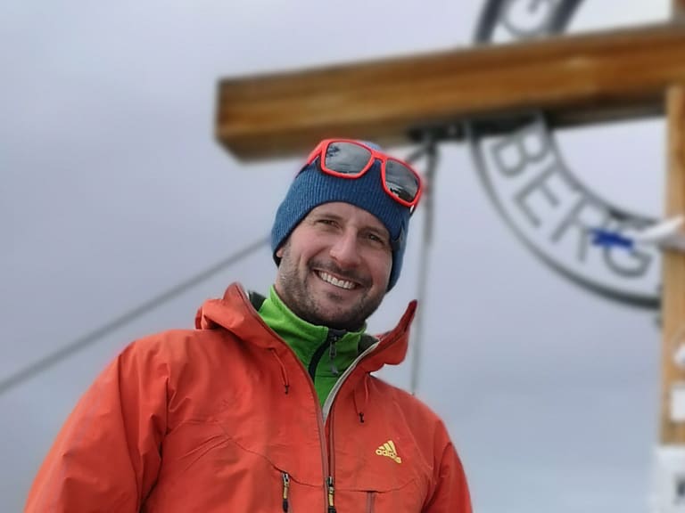 Am Gipfelkreuz mit bunten Gebetsfähnchen steht Bergführer Andreas Strasser in oranger Softshelljacke im dichten Nebel.