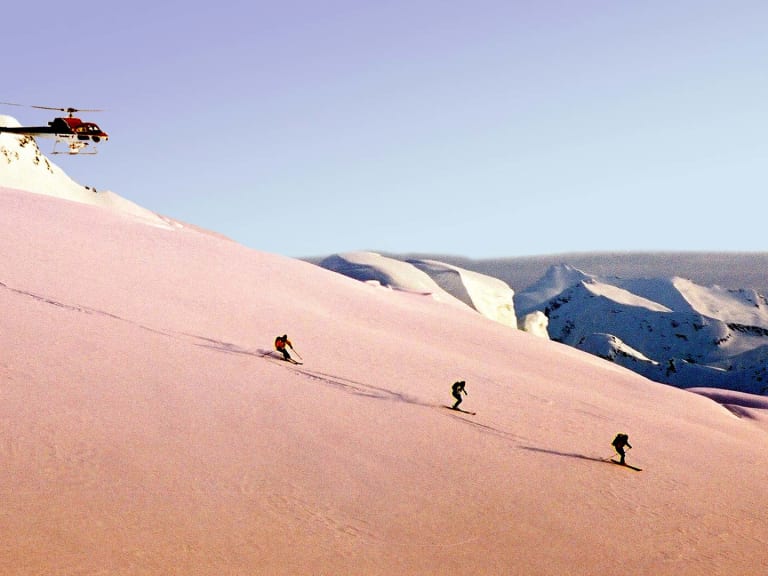 Bergführer Hubert Walther begleitet drei Skifahrer auf unverspurtem Schneehang, während rot-schwarzer Hubschrauber über ihnen schwebt.