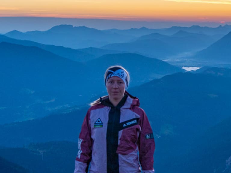 Bergwanderführerin Maria Müller steht auf Gipfelplateau mit Kalksteinblock vor weitläufigem Alpenpanorama bei goldener Lichtstimmung.