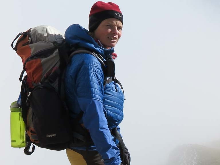 Mit blauer Daunenjacke und roter Mütze steht Bergwanderführerin Andrea Beuchlen auf einem felsigen Grat vor einer Berghütte.