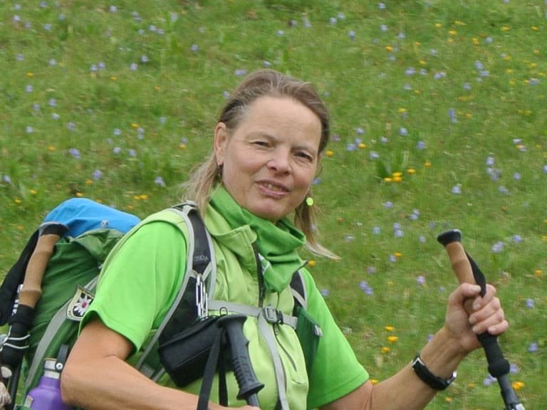 In blühender Almwiese mit gelben und violetten Wildblumen steht Bergwanderführerin Andrea Beuchlen mit grünem Rucksack und Wanderstöcken.